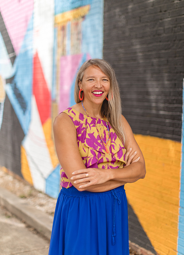 Smiling woman with her arms crossed, standing in front of a colorfully painted brick wall.