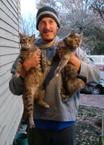 Man with beanie holding two cats.